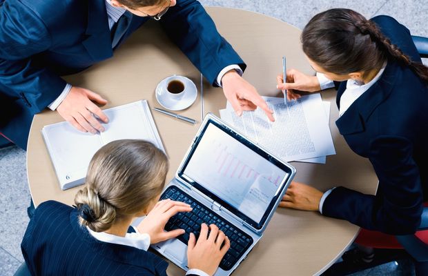 Seen from above, three colleagues work at a round table with a laptop and papers