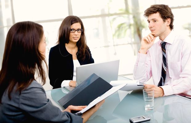 Three young colleagues sit at a glass table with a laptop and a folder