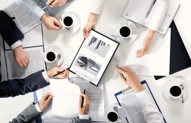Seen from above, hands with pens over printed charts and coffee cups on a table